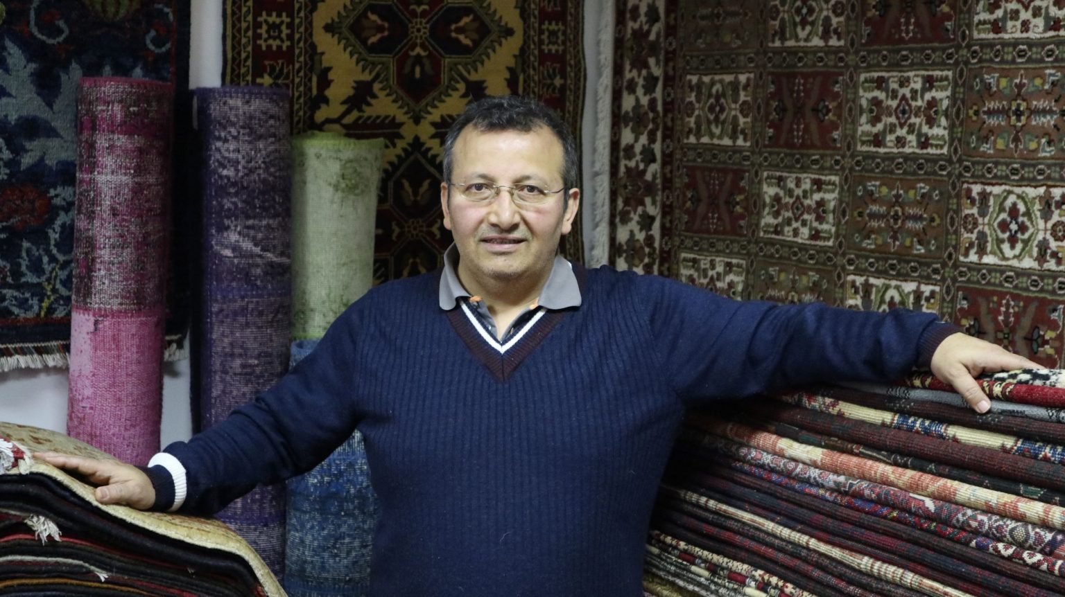 Persian carpet expert standing among stacked handmade rugs in a showroom.